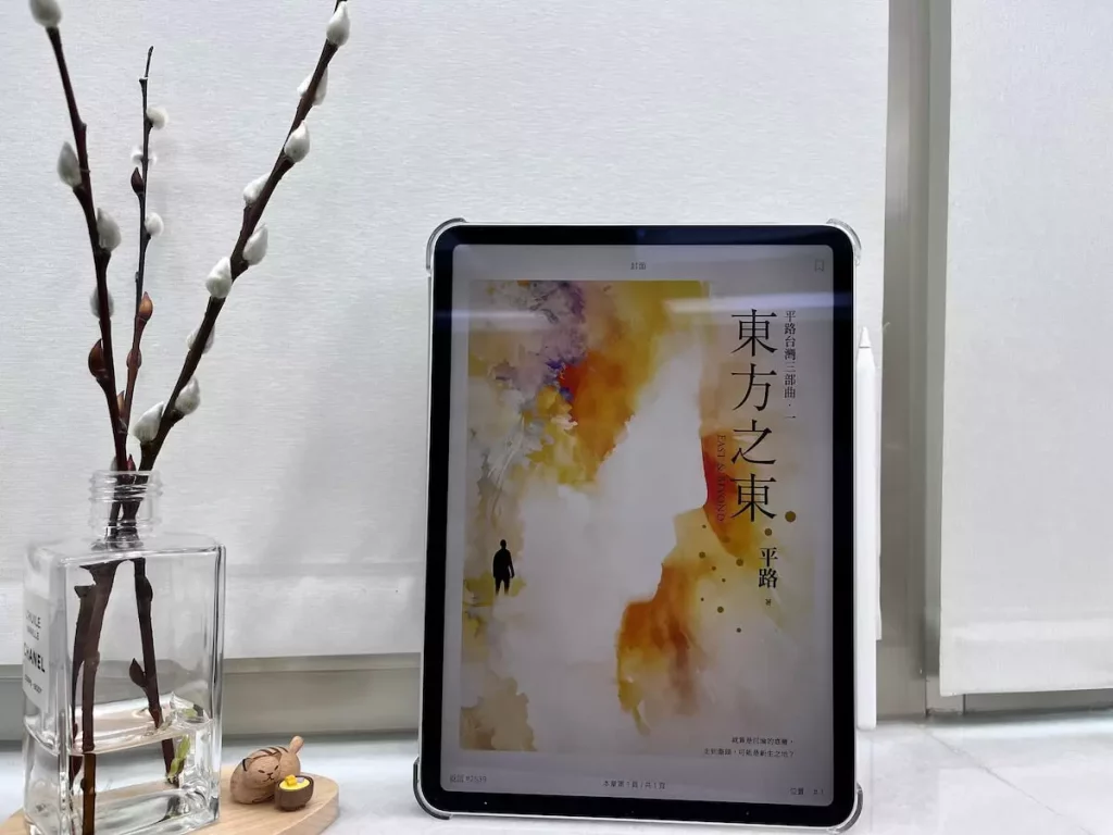 這是一台立在窗邊白色大理石窗台上的iPad Pro，螢幕顯示電子書東方之東的封面，平板旁邊有一個玻璃瓶，瓶中有水，還有3枝銀柳置放在木質底座上