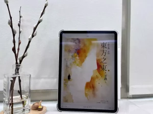 這是一台立在窗邊白色大理石窗台上的iPad Pro，螢幕顯示電子書東方之東的封面，平板旁邊有一個玻璃瓶，瓶中有水，還有3枝銀柳置放在木質底座上