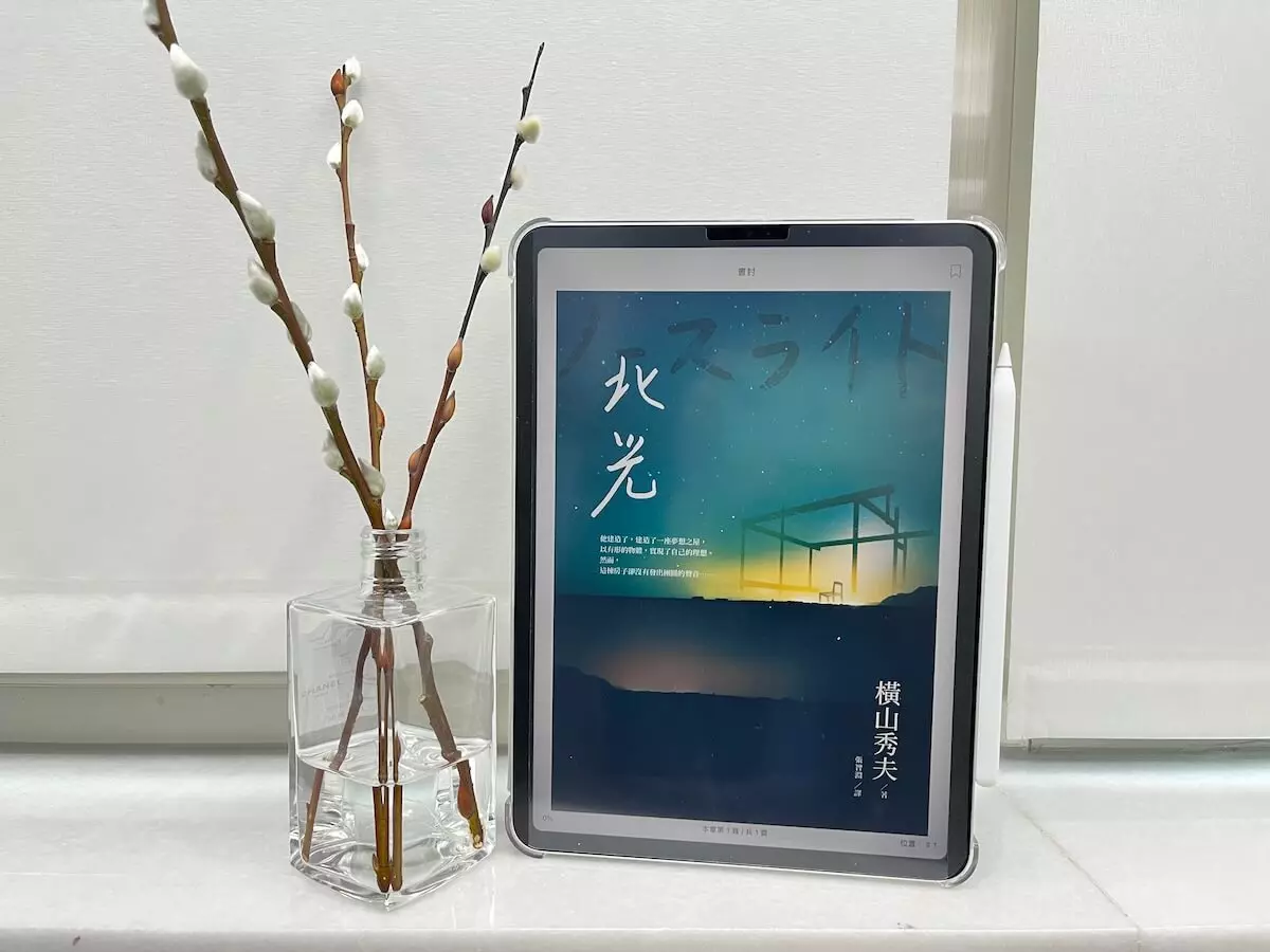 這是一台立在窗邊白色大理石窗台上的iPad Pro，螢幕顯示電子書北光小說的繁體中文版封面，平板旁邊有一個玻璃瓶，瓶中有水，還有3枝銀柳