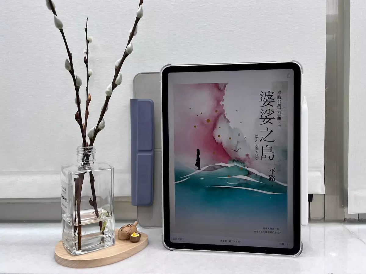 這是一台立在窗邊白色大理石窗台上的iPad Pro，螢幕顯示電子書婆娑之島的封面，平板旁邊有一個玻璃瓶，瓶中有水，還有3枝銀柳置放在木質底座上