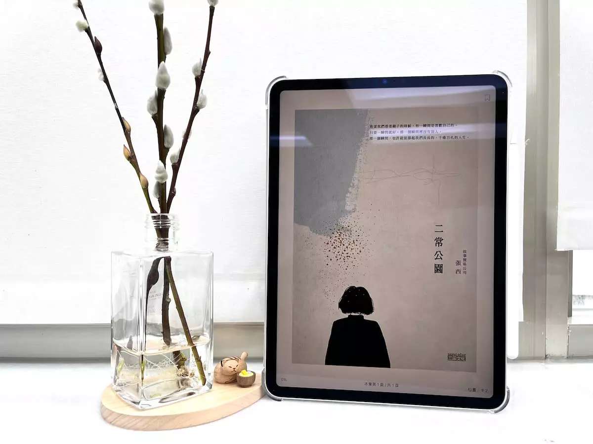 這是一台立在窗邊白色大理石窗台上的iPad Pro，螢幕顯示電子書二常公園的封面，平板旁邊有一個玻璃瓶，瓶中有水，還有3枝銀柳置放在木質底座上