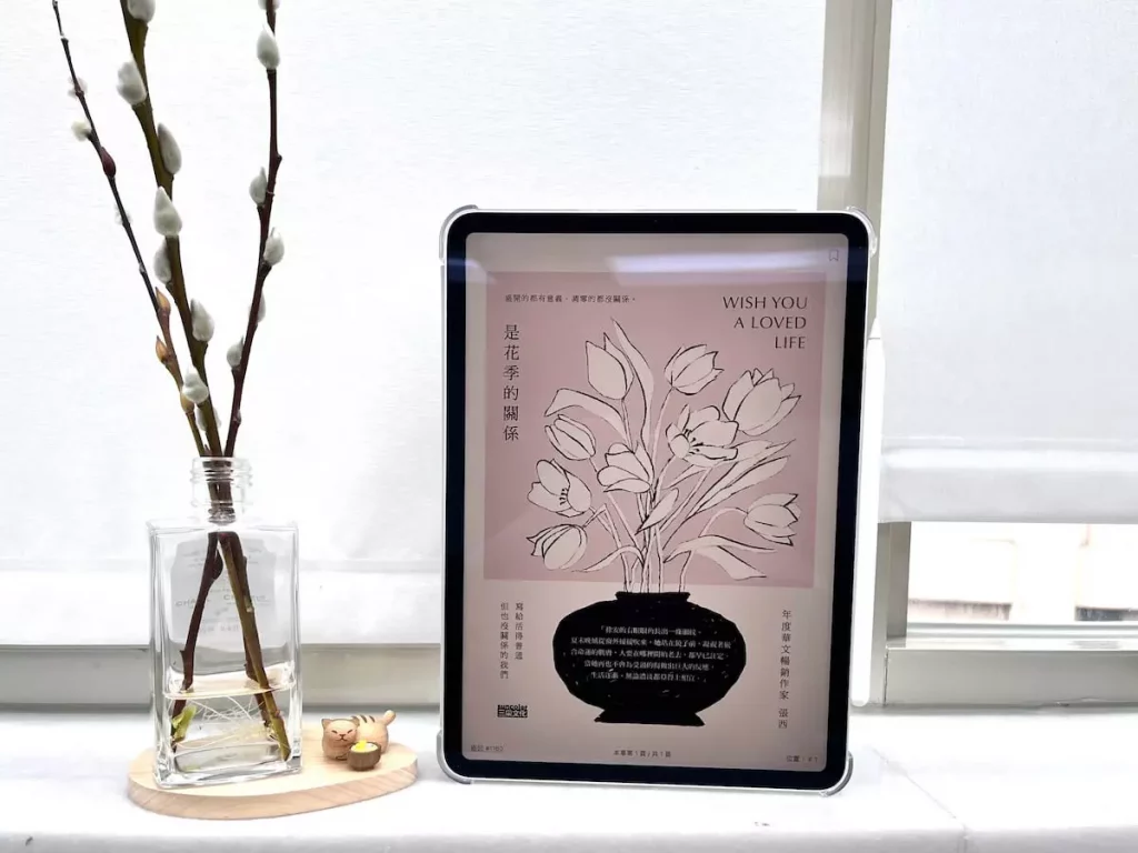 這是一台立在窗邊白色大理石窗台上的iPad Pro，螢幕顯示電子書是花季的關係的封面，平板旁邊有一個玻璃瓶，瓶中有水，還有3枝銀柳置放在木質底座上