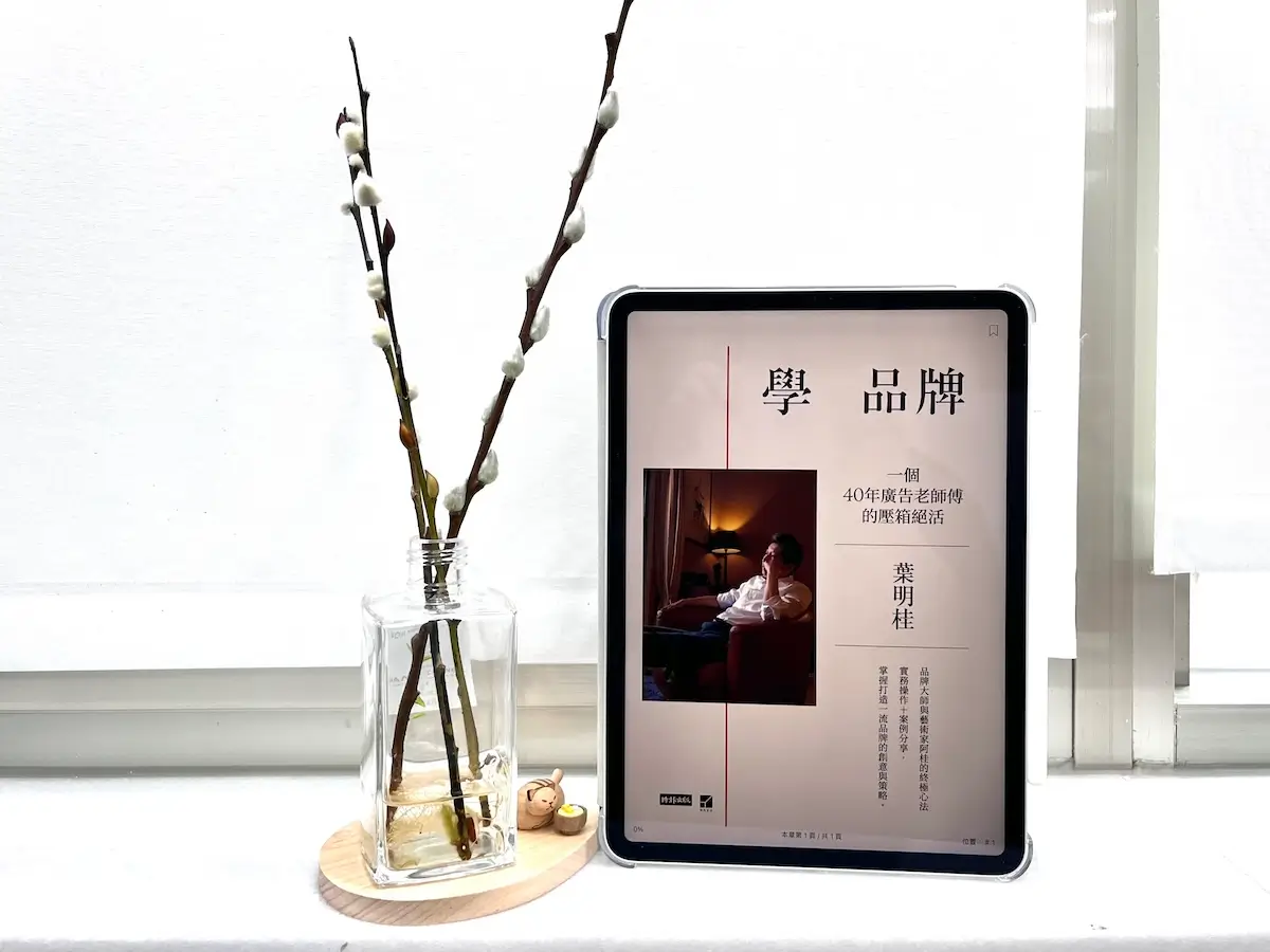 這是一台立在窗邊白色大理石窗台上的iPad Pro，螢幕顯示電子書學品牌的封面，平板旁邊有一個玻璃瓶，瓶中有水，還有3枝銀柳置放在木質底座上