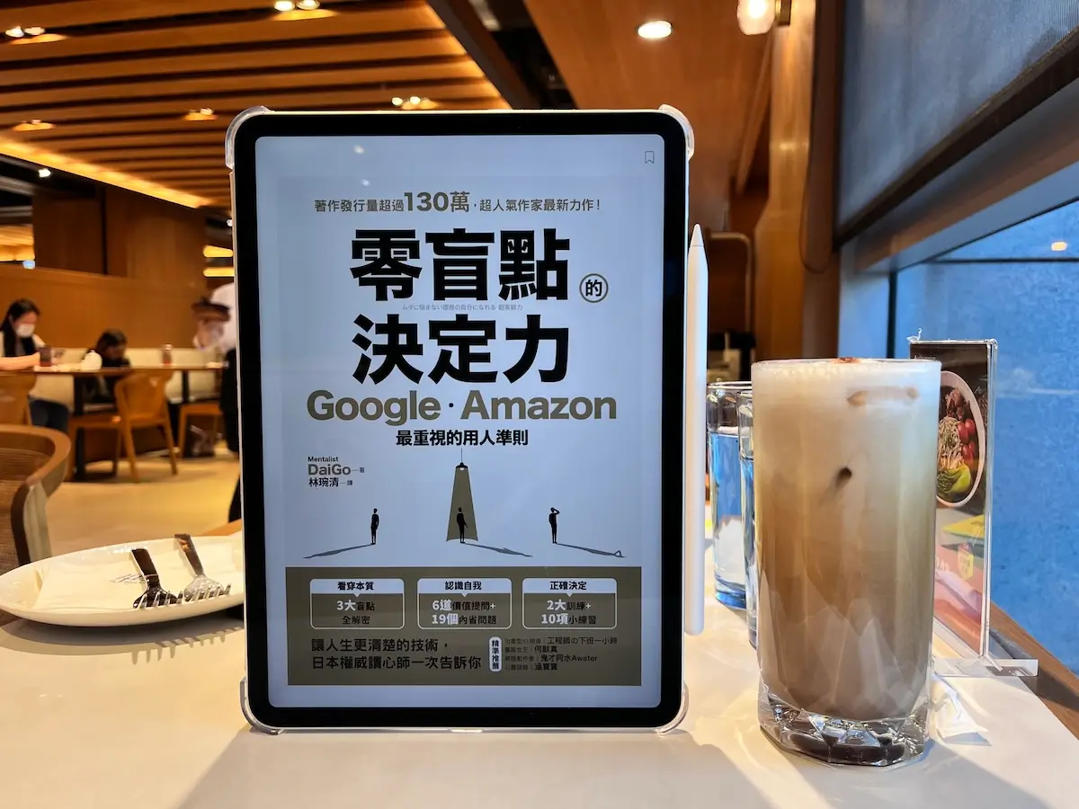 在靠窗邊的咖啡廳桌上立有一台iPad顯示零盲點的決定力電子書封面，iPad旁有一杯冰磨卡加啡玻璃杯