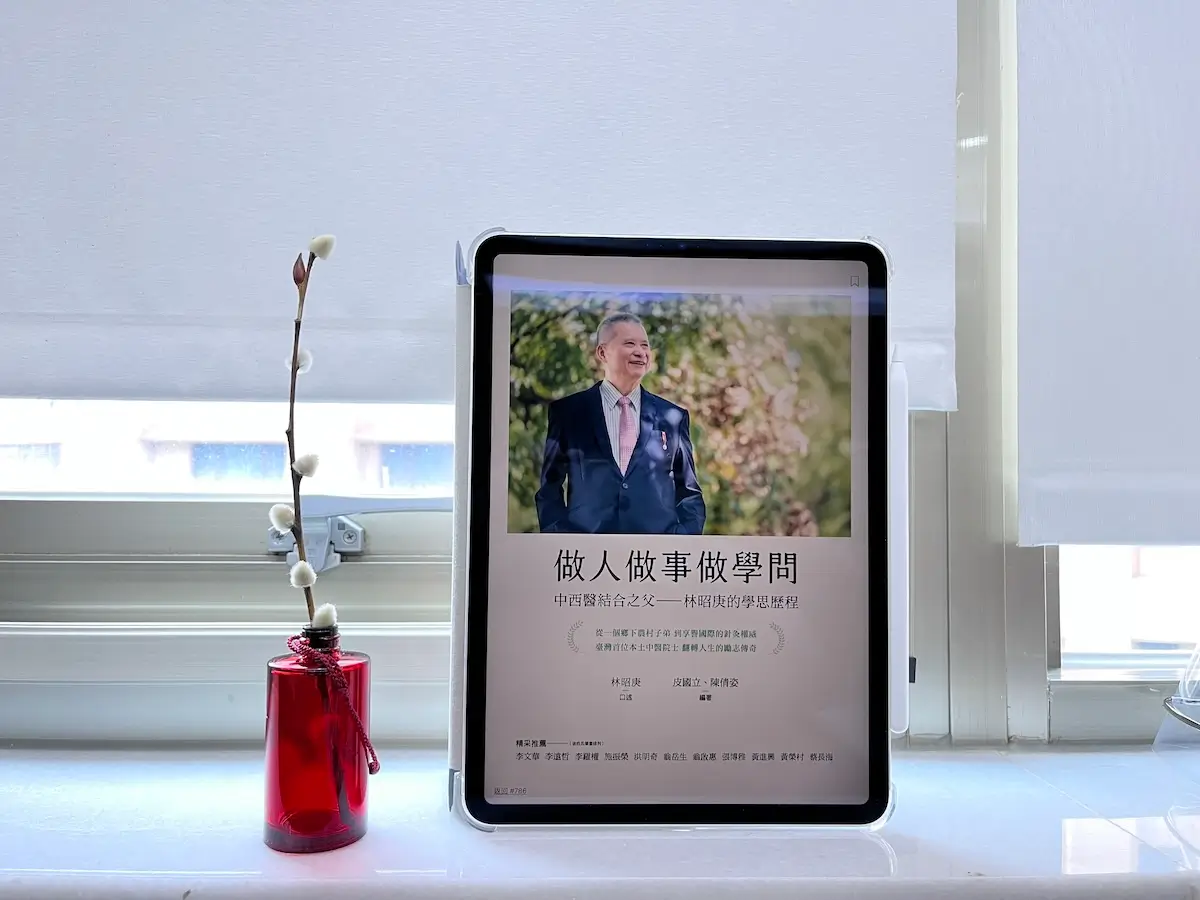 這是一台立在窗邊白色大理石窗台上的iPad Pro，螢幕顯示電子書做人做事做學問：中西醫結合之父——林昭庚的學思歷程的封面，平板旁邊有一個紅色玻璃瓶，插有1枝銀柳