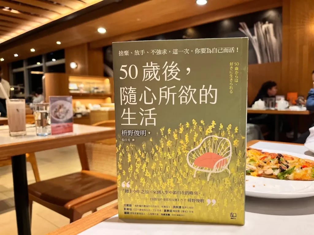 50歲後，隨心所欲的生活 繁體中文版的紙本書立於咖啡廳木桌上，書後還有有一片薄餅放在瓷盤上