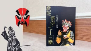 這是一本 戲班悟空七十三變 書本放在貼皮木紋桌面上，旁邊設計京劇臉譜很女伶的圖示點綴圖片