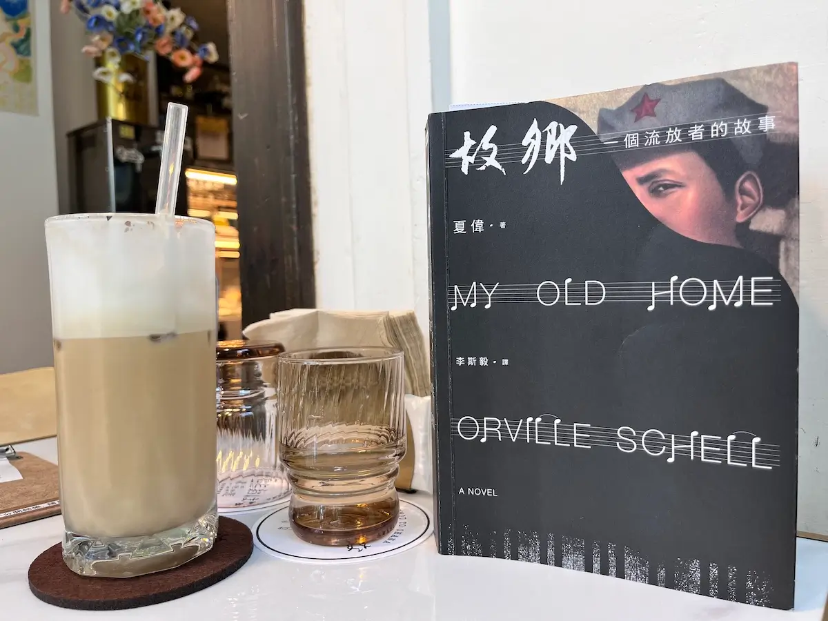 故鄉：一個流亡者的故事 繁體中文版的紙本書立於咖啡廳桌上，書本旁有一個玻璃水杯，內約半杯水，水杯旁有一杯冰拿鐵咖啡，杯內有透明玻璃吸管 ，杯底有深咖啡色圓形杯墊