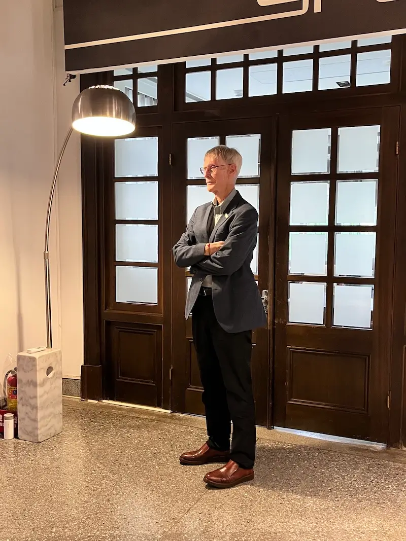 故鄉:一個流亡者的故事 的作者夏偉雙手叉在胸腹間站立於碎石面地板上供記者拍照,旁有一燈具亮者微黃燈