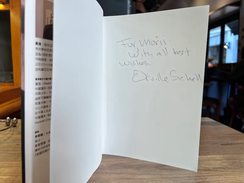 故鄉:一個流亡者的故事 繁體中文版的紙本書立於咖啡廳木桌上,內頁有作者親簽的簽名,上有文字For Morii: With all best wishes. Orville Schell