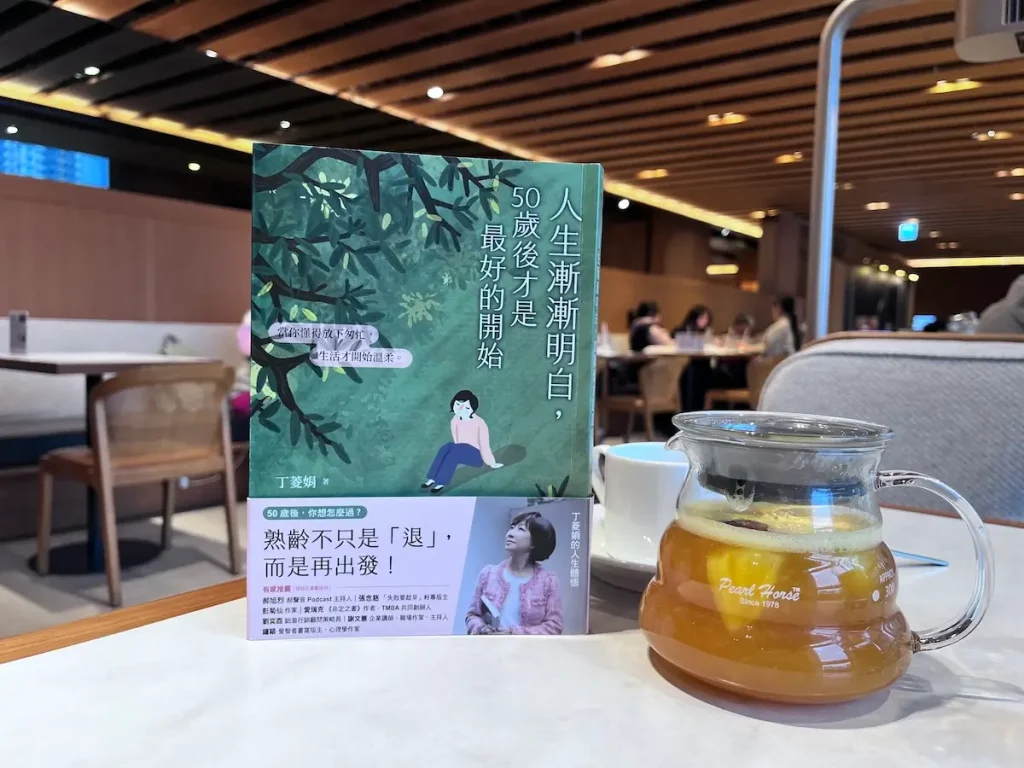 人生漸漸明白，50歲後才是最好的開始 繁體中文版的紙本書立於咖啡廳木桌上，書旁有一壺水果茶