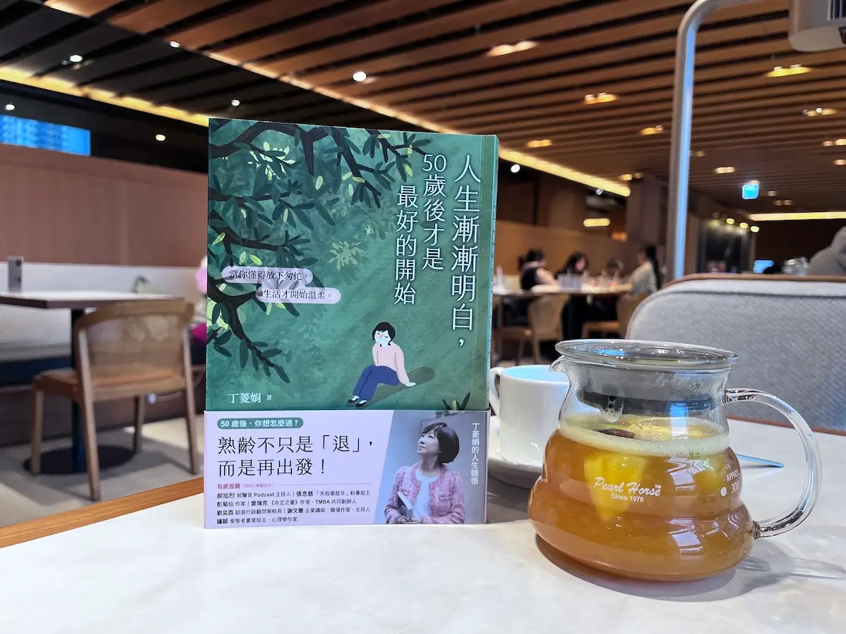 人生漸漸明白，50歲後才是最好的開始 繁體中文版的紙本書立於咖啡廳木桌上，書旁有一壺水果茶